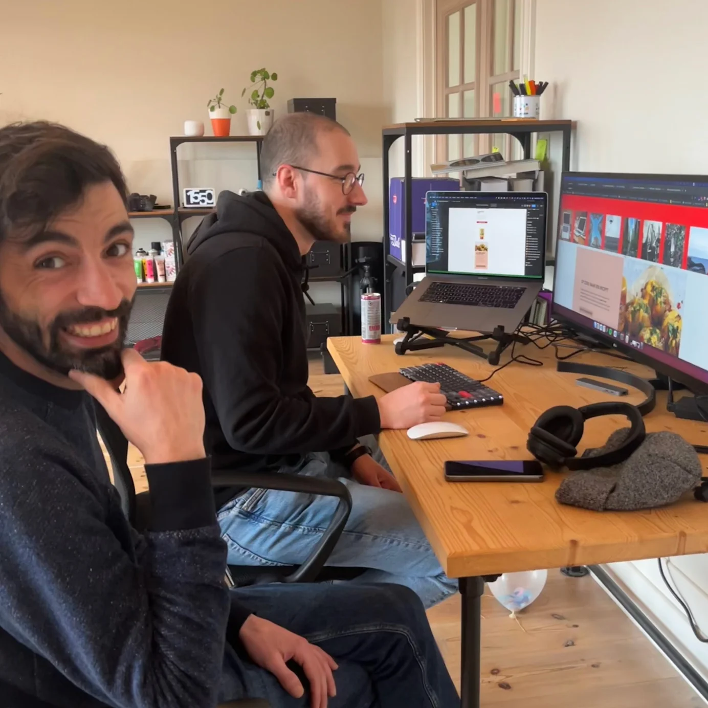 Two guys working together at one desk, one guy is looking at the screen, the other is looking at the camera